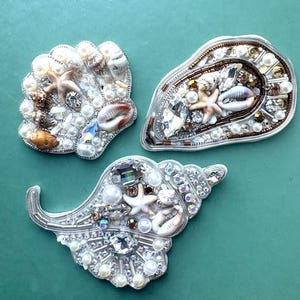 May include: Three decorative shell-shaped brooches embellished with white pearls, small natural seashells, tiny starfish, and clear faceted crystals. Each piece features intricate beadwork and metallic thread detailing, creating a textured, multi-dimensional appearance on each shell form.