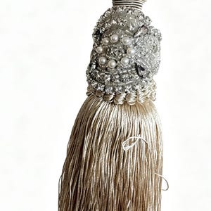 May include: A decorative tassel with a silver and pearl beaded top and a long, flowing beige fringe. The top is intricately detailed with rhinestones and small pearls, and the fringe is made of fine, silky threads.
