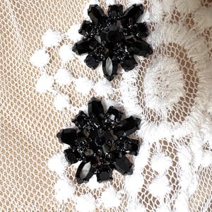 Puede incluir: Dos adornos florales negros con detalles facetados en forma de joya. Los adornos se colocan sobre un fondo de encaje blanco con un diseño calado delicado. Los acentos negros crean un contraste llamativo.