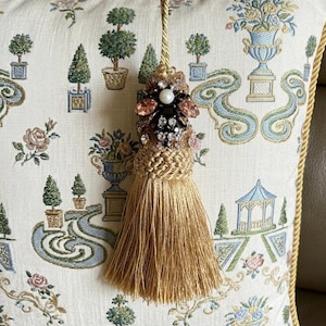 May include: A decorative gold tassel featuring a beaded ornament with a central white pearl, pink and black crystals, and a braided cord. The tassel is positioned against a cream-coloured pillow with a blue, green, and pink garden-themed pattern.