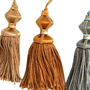 May include: Three decorative tassels in gold, orange, and grey. Each tassel has a bulbous top with a diamond-shaped centre and a long, braided fringe. These tassels are designed for home decor.
