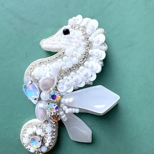Puede incluir: Un broche con forma de caballito de mar elaborado con cuentas blancas, lentejuelas y cristales iridiscentes. El diseño presenta una cuenta negra para el ojo, detalles en hilo metálico plateado y dos formas blancas translúcidas.