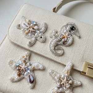 Può includere: Una borsa strutturata color crema con quattro intricate applicazioni di perline. I disegni includono una tartaruga marina, un cavalluccio marino e due stelle marine, decorati con perle bianche, piccole conchiglie, cristalli e filo d'argento.