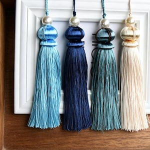 Peut inclure: Quatre pompons décoratifs dans des tons de bleu et crème, chacun avec une perle en haut. Les pompons sont disposés devant un cadre photo blanc, mettant en valeur leurs longueurs et textures variées. Les pompons sont en matière soyeuse.