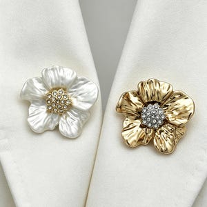 Puede incluir: Dos broches con forma de flor. Uno es blanco con un centro dorado, y el otro es dorado con un centro plateado. Ambos broches están sobre un fondo de tela blanca.