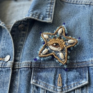 Op de afbeelding: Een stervormige broche met ingewikkeld kraalwerk en borduurwerk. Het midden toont een gestileerd oogmotief in gouden, witte en zwarte draden. De ster is versierd met witte ovale parels, lichtblauwe kristallen en donkerblauwe kralen.