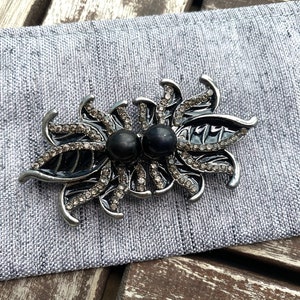 May include: A silver brooch with black enamel and rhinestones in the shape of two stylised flowers. The brooch has two black pearl-like accents in the centre.