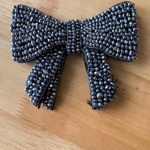 K&ouml;nnte beinhalten: Ein schleifenf&ouml;rmiges Accessoire, das mit dunkelgrauen, facettierten Perlen bedeckt ist. Die Schleife besteht aus einem Mittelknoten und zwei herabh&auml;ngenden Enden, die dicht mit reflektierenden, metallisch wirkenden Perlen besetzt sind.