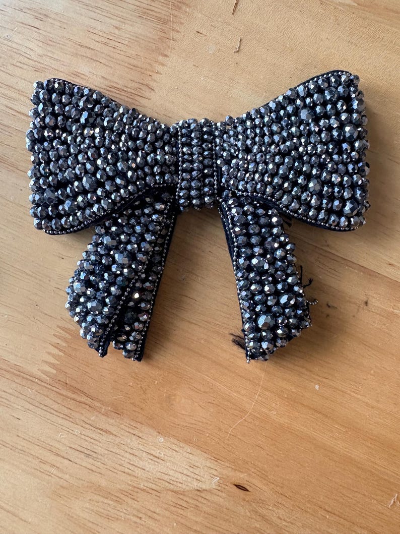 K&ouml;nnte beinhalten: Ein schleifenf&ouml;rmiges Accessoire, das mit dunkelgrauen, facettierten Perlen bedeckt ist. Die Schleife besteht aus einem Mittelknoten und zwei herabh&auml;ngenden Enden, die dicht mit reflektierenden, metallisch wirkenden Perlen besetzt sind.
