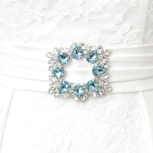Puede incluir: Un broche decorativo con una disposición circular de cristales azul claro en forma de pera, rodeados por un borde de cristales redondos más pequeños y transparentes. El broche está centrado sobre una faja de satén blanco y encaje blanco.