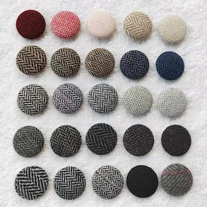 May include: A collection of twenty-five circular fabric-covered buttons arranged in a five-by-five grid. The buttons feature various colors including red, beige, brown, blue, gray, and black, with most displaying a woven herringbone pattern.