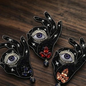 May include: Three black hand-shaped brooches featuring embroidered star patterns, a central realistic eye with a purple iris, and dangling teardrop-shaped crystals in red, blue, and orange hues, accented with small black beads and silver thread.