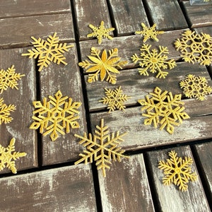 Puede incluir: Una colección de 14 adornos de copo de nieve dorados sobre una superficie de madera. Los copos de nieve son de varios tamaños y estilos.