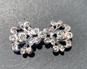 May include: A silver bow-shaped brooch with clear rhinestones. The brooch has a delicate, intricate design.