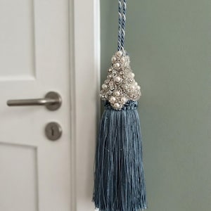 May include: A decorative curtain tassel featuring a long, dusty blue fringe suspended from a braided cord. The top section is embellished with a cluster of white faux pearls and clear faceted crystals set in a silver-toned metal frame.