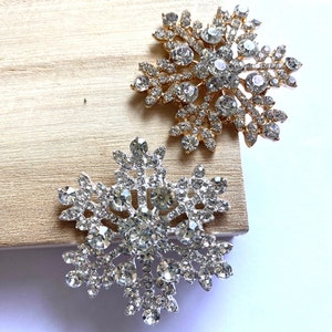 May include: Two snowflake brooches, one silver and one gold, with clear rhinestones. The brooches are on a wooden surface.