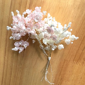 Puede incluir: Un delicado accesorio floral para el cabello con racimos de flores rosas y blancas con detalles de perlas. Las flores están cableadas con tallos dorados y plateados, atadas en la base. El arreglo descansa sobre una superficie de madera.