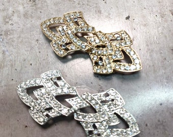 May include: Two ornate brooches with a geometric design, one gold and one silver, both encrusted with clear rhinestones.