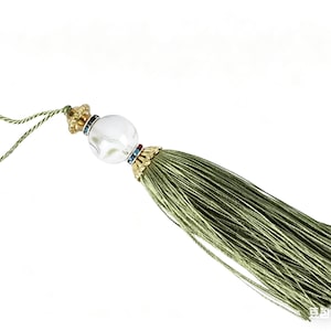 May include: An olive green tassel with a decorative top. The top features a clear, round bead with gold and jewel accents. The tassel is made of many strands of olive green thread and is attached to a thin, twisted cord.