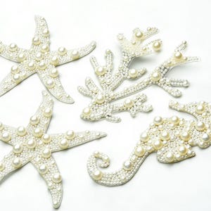Puede incluir: Adornos decorativos con temática marina, que incluyen dos estrellas de mar, una rama de coral y un caballito de mar. Cada pieza está cubierta de pequeñas cuentas blancas y perlas redondas más grandes de color crema, creando un efecto brillante. Sobre fondo blanco.