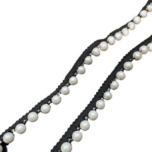 May include: Black beaded trim with white faux pearls. The trim features a black woven base with evenly spaced, round, white pearls. The pearls are attached with small silver-colored beads. This trim is suitable for various craft and sewing projects.