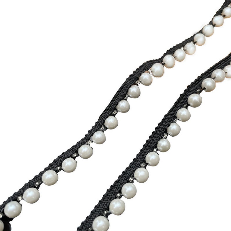 May include: Black beaded trim with white faux pearls. The trim features a black woven base with evenly spaced, round, white pearls. The pearls are attached with small silver-colored beads. This trim is suitable for various craft and sewing projects.