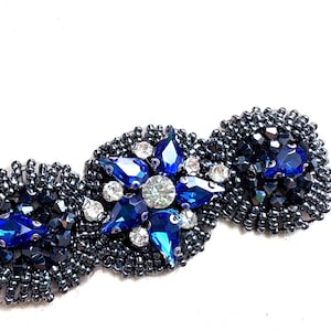 以下が含まれることがあります： 3つのセクションが繋がった、装飾的なビーズ刺繍。各セクションは小さな濃い色のビーズで密に覆われています。中央のセクションは、青とクリアのクリスタルを使ったスターバーストのデザインで、他のセクションには青いアクセントがあります。