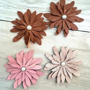May include: Four flower-shaped embellishments made of textured faux leather. Two flowers are brown, one is light pink, and one is a pale beige. Each flower features multiple layered petals radiating from a central white pearl-like bead accent.