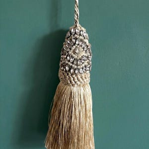 May include: A decorative tassel featuring a beige cord and a dense fringe of matching beige threads. The upper section is embellished with a cluster of clear faceted crystals and small white pearls, secured by a braided beige rope collar.