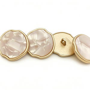 Op de afbeelding: Vier decoratieve knopen met een parelmoer, gebroken witte kern en een goudkleurige, golvende rand. De achterkanten van de knopen zijn goudkleurig met een lus om op te naaien. De knopen zijn ideaal voor naaiprojecten.
