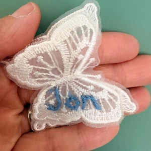 Puede incluir: Parche de mariposa bordada en blanco con detalles delicados en las alas. La palabra "Don" está bordada en azul en el ala inferior. El parche se sostiene en una mano sobre un fondo turquesa.