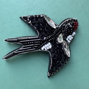 Puede incluir: Un broche con forma de golondrina elaborado con lentejuelas negras, cuentas de vidrio negras y cuentas de semillas blancas. El ave presenta cristales facetados transparentes en las alas y un pequeño grupo de cuentas rojas en el pico, bordeado con hilo plateado.