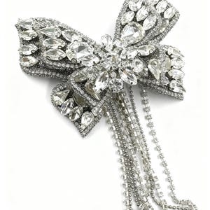 May include: A silver-toned bow-shaped brooch, heavily embellished with clear, sparkling rhinestones. The brooch features a central cluster of larger stones and cascading chains of smaller rhinestones, creating a glamorous, eye-catching accessory.