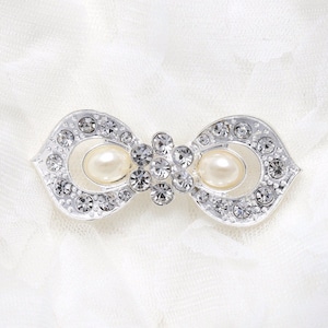 Puede incluir: Un broche de plata en forma de lazo con brillantes transparentes y dos falsos acentos de perla.