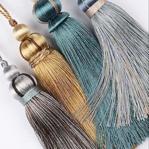 May include: Close-up of four decorative tassels in shades of gold, teal, and grey. Each tassel features a beaded top and a long, flowing fringe. The tassels are designed for home decor and add a touch of elegance.