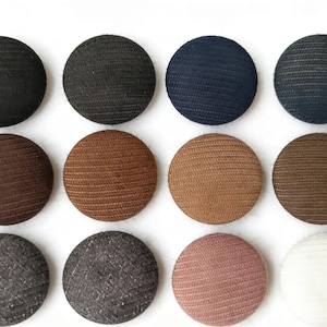 May include: Twelve round buttons in a variety of colours, including black, navy, brown, and pink. The buttons have a textured, woven appearance and are arranged on a white background.