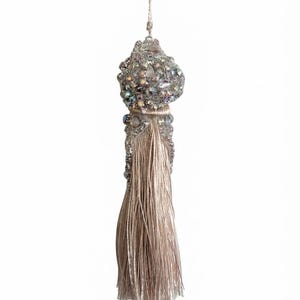 May include: A decorative tassel featuring a beige silk-like fringe suspended from a top section heavily embellished with iridescent, clear, and silver-toned beads and crystals. A thin matching cord extends from the top for hanging purposes.