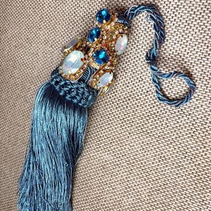 May include: A decorative tassel featuring a blue cord and a dense fringe of blue threads. The upper section is embellished with gold-tone metal settings holding round blue crystals and oval iridescent white stones, finished with a twisted blue loop.