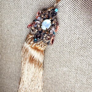 May include: A decorative tassel with a beaded top and a gold cord. The top features an oval, light blue stone surrounded by various colourful gems. The tassel is made of gold-coloured threads.