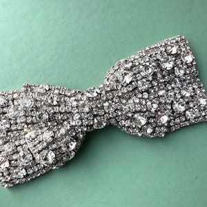 May include: A silver bow-shaped brooch made entirely of rhinestones. The brooch is a sparkly accessory that can be used to add a touch of glamour to any outfit.