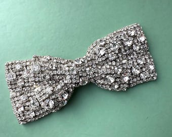 May include: A silver bow-shaped brooch made entirely of rhinestones. The brooch is a sparkly accessory that can be used to add a touch of glamour to any outfit.