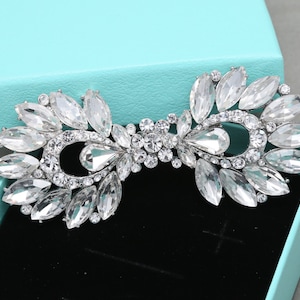 Puede incluir: Un broche de plata con un diseño grande y ornamentado con diamantes de imitación transparentes. El broche tiene forma de lazo con dos grandes diamantes de imitación en forma de lágrima en el centro.