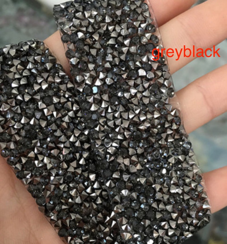 May include: Close-up of a roll of grey-black rhinestone trim. The trim is made of small, pointed rhinestones that are closely spaced together.
