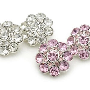 May include: Close-up of three flower-shaped rhinestone buttons. Two are clear, and one is pink. Each button has a silver-colored metal base and is composed of multiple rhinestones of varying sizes.
