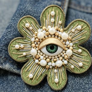 May include: A flower-shaped brooch featuring a central realistic green eye surrounded by white and gold beads, pearls, and metallic silver and gold thread embroidery on a sage green fabric base with a stitched border detail.
