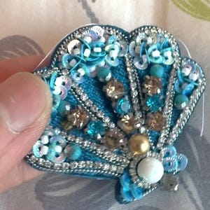 Puede incluir: Un broche con forma de concha que presenta un bordado intrincado en tonos azules. El diseño está adornado con lentejuelas iridiscentes, hilo metálico plateado, cuentas azul claro, cristales facetados color champán y una perla blanca central.