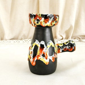 May include: A black ceramic vase with a handle and a colorful abstract design in orange, yellow, and white. The vase has a wide mouth and a narrow base.