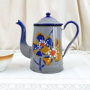 Pode incluir: Uma chaleira de esmalte cinza com uma borda azul e um desenho floral em amarelo, azul, branco e vermelho. A chaleira tem uma tampa e uma alça. Também há uma tigela branca com uma borda marrom na imagem.