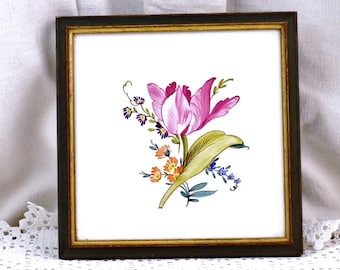 May include: A framed tile featuring a hand-painted floral design. The tile depicts a pink and white tulip with green leaves and other smaller flowers in shades of blue, purple, and orange.
