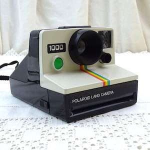 Puede incluir: Una cámara Polaroid Land Camera 1000 vintage, con un cuerpo negro y crema y una franja de arcoíris. La cámara tiene una lente negra y un botón verde. Se adjunta una correa negra.